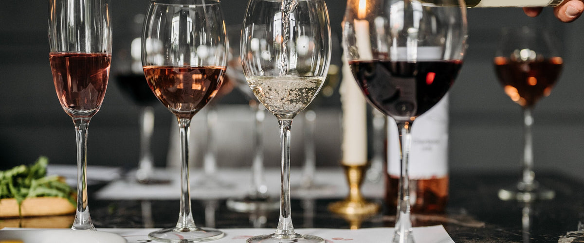 Unsere Weinwelt: Gedeckter Tisch mit Rosé-, Weiß- und Rotweingläsern, es wird gerade eingeschenkt, Kerze im Hintergrund