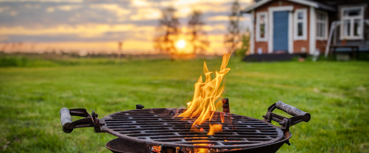 Grillweine: Feuerschale/Grill mit Flammen auf dem Rasen, Holzhaus und Sonnenuntergang dahinter