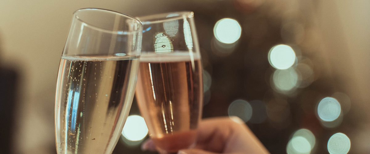 Jubiläum: Zwei Gläser Sekt und Rosé-Sekt stoßen an, warme Lichtkugeln dahinter