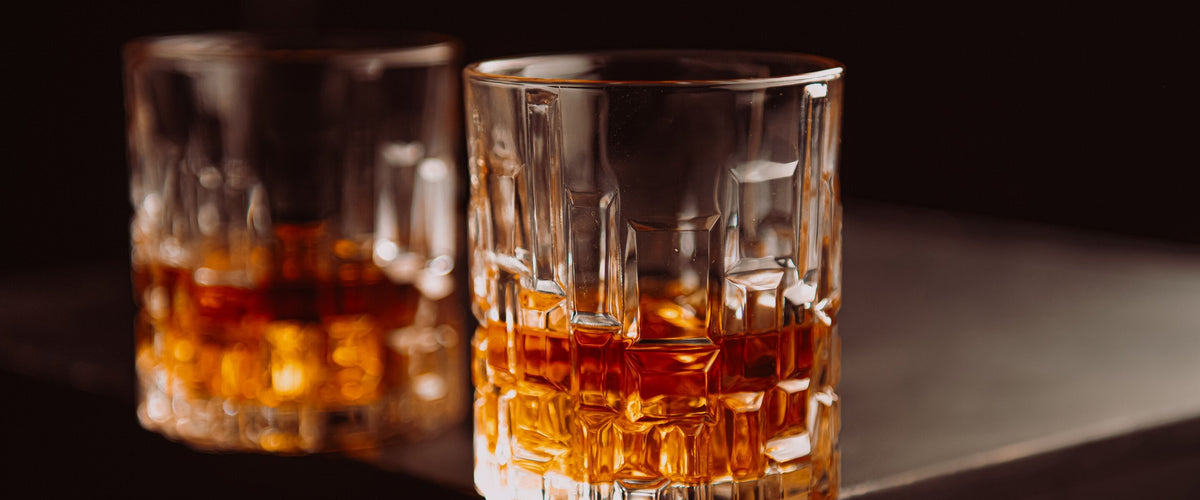 Spirituose: Zwei Kristall-Tumbler mit bernsteinfarbener Spirituose vor dunklem Grund, seitliches Licht