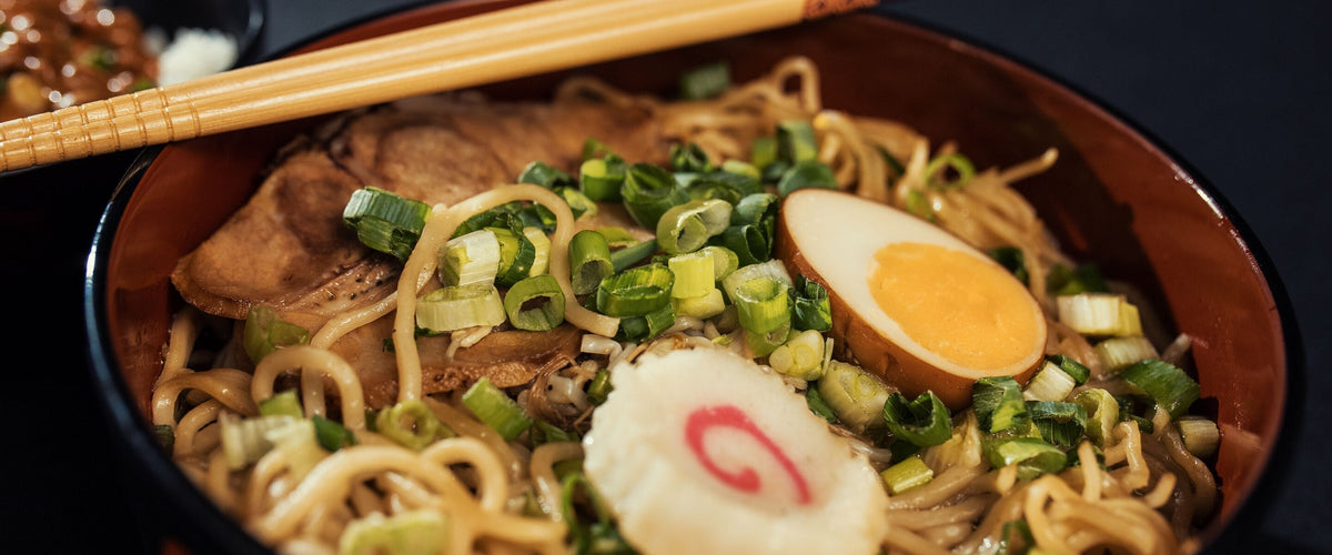 Wein zur asiatischen Küche: Ramen mit Ei, Lauchzwiebeln und Fleisch in dunkler Schale, Stäbchen darüber