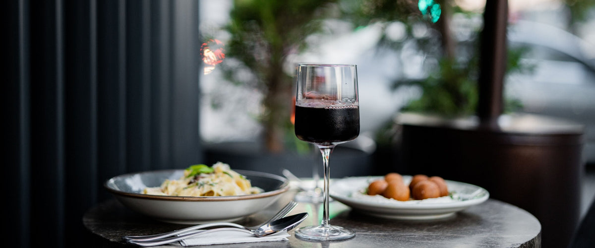 Wein zu Pilzen & Trüffel: Glas Rotwein auf einem Bistrotisch mit einem Teller Pasta und einem Teller Kroketten