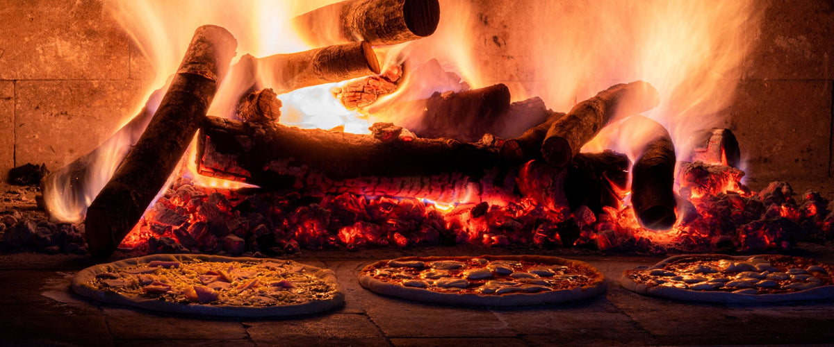 Wein zu Pizza: Brennende Holzscheite im Steinofen, davor drei Pizzen auf dem Ofenboden