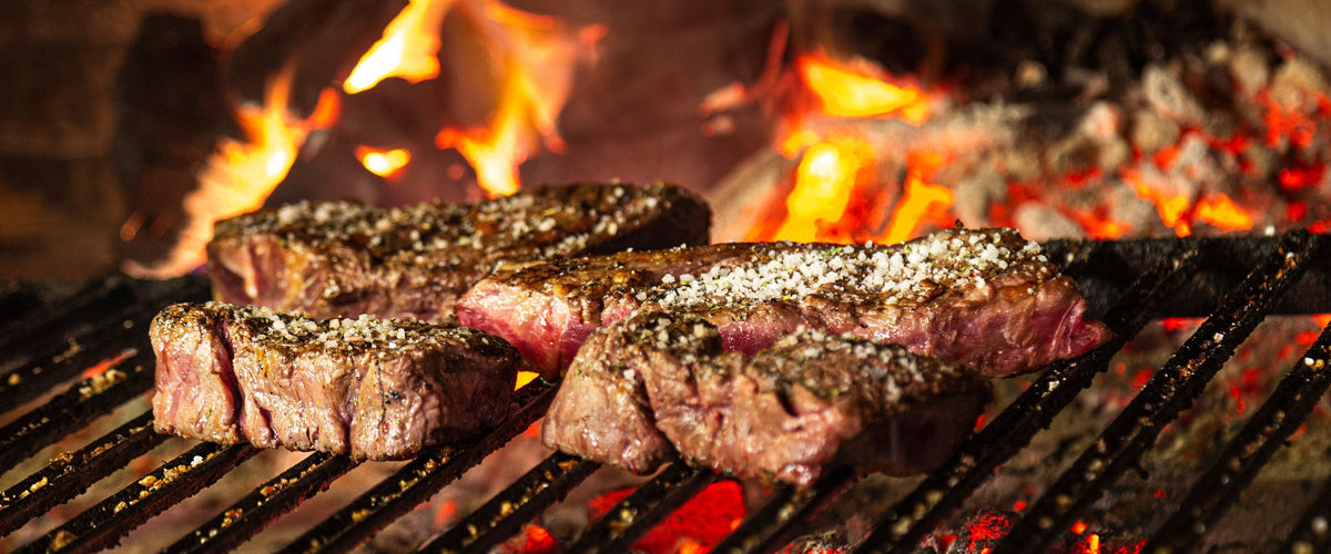 Wein zu Rind & Steak: Drei gewürzte Steaks auf dem Rost, dahinter loderndes Kaminfeuer und Glut