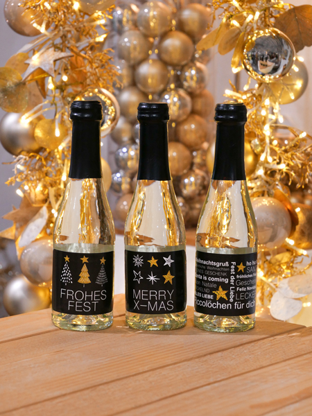Drei Piccolo-Flaschen Perlwein von Vom Feinsten mit den Etiketten Frohes Fest, Merry X-Mas und Weihnachtsgruß vor goldenen Christbaumkugeln