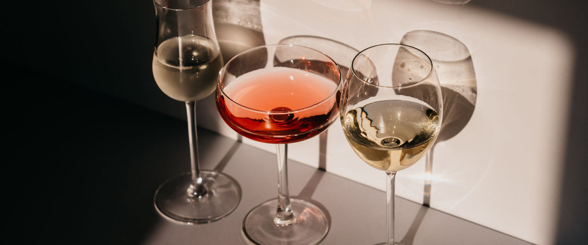 Alkoholfreie Weine & Alternativen: Drei Stielgläser mit hellem und roséfarbenem Getränk in weichem Streiflicht auf dem Tisch