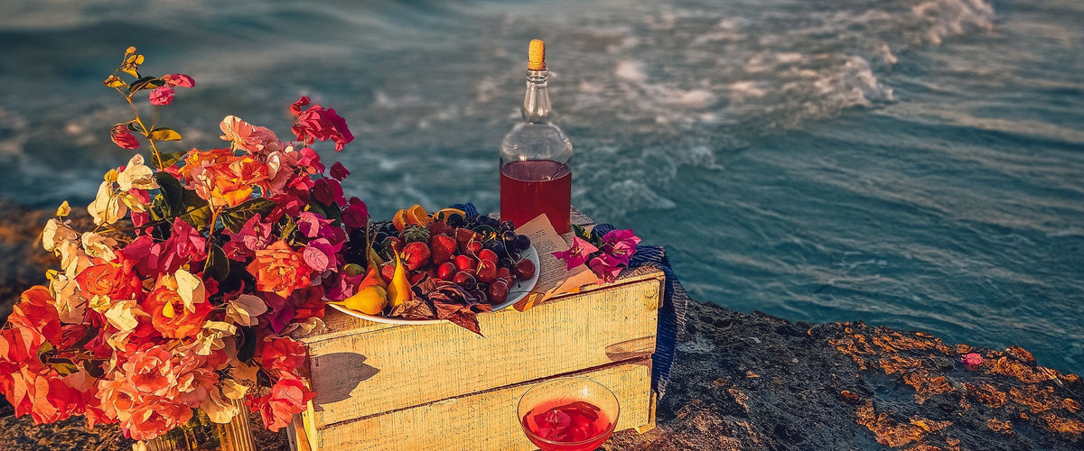 Wein zur mediterranen Küche: Felsen am Meer im Abendlicht, Holzkiste mit Bougainvillea und Obst, Flasche und Glas Rosé