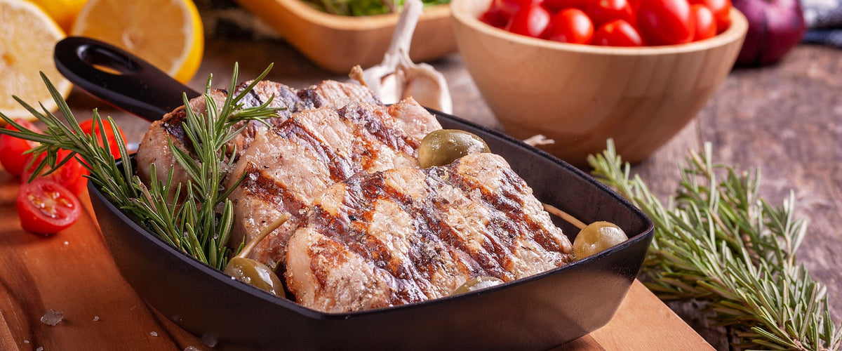 Wein zu Schwein: Gegrillter Braten in der gusseisernen Pfanne mit Rosmarin und Oliven auf Holz, Tomaten dahinter