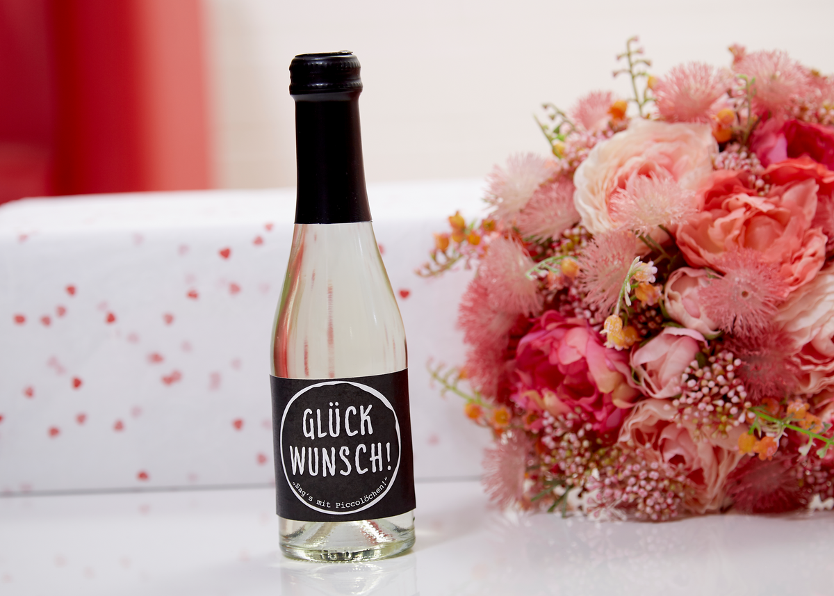 Piccolo-Flasche Perlwein Glückwunsch von Vom Feinsten neben einem Blumenstrauß und einem Geschenk