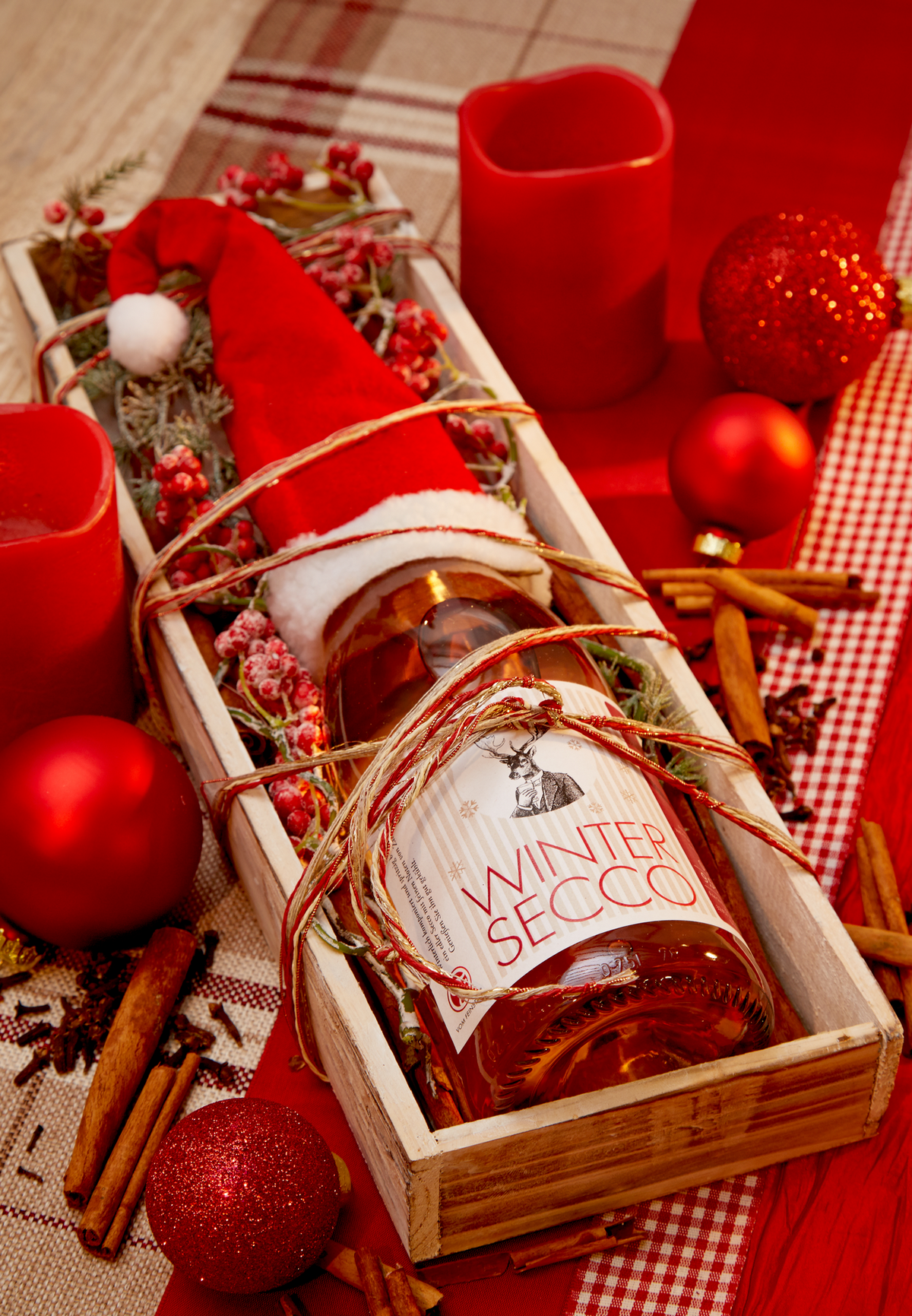 Flasche Wintersecco mit Zimt und Nelke von Vom Feinsten mit Weihnachtsmütze in einer Holzkiste mit Zimtstangen und roten Kerzen