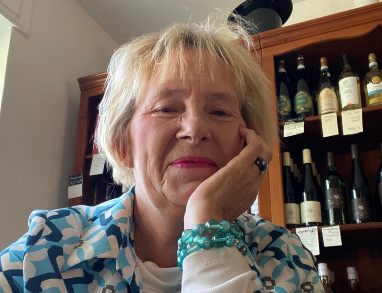 Elke Bastert im Weinkontor in Bad Oeynhausen, das Kinn auf die Hand gestützt, hinter ihr das Weinregal